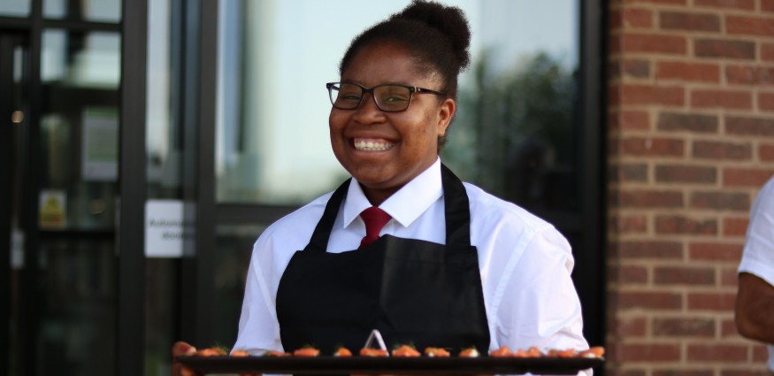 A waitress wearing an apron over a shirt and tie carries a food platter.